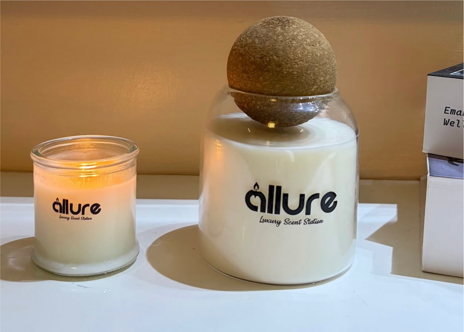 Allure branded candles in glass vessels, one lit, on a sunlit ledge